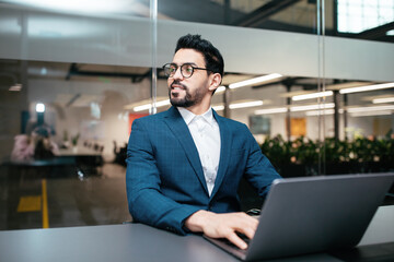 Serious busy attractive millennial muslim businessman with beard in suit, glasses works with pc in office