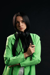 Elegant pretty girl looking at camera with sad expression inside. Portrait view of thoughtful woman in green blazer with headphones on neck, isolated on black studio background. Concept of emotions.