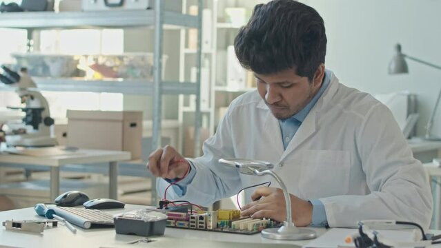 Medium slowmo of Biracial male engineer in white coat fixing motherboard in laboratory while his colleague inspecting pcb in background
