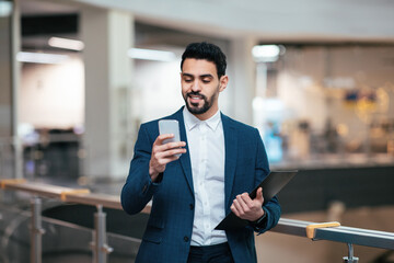 Cheerful handsome millennial arabic businessman with beard in suit with tablet looks at smartphone...