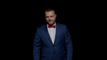 An elegant man in a blue tuxedo and a red tie smiles sweetly against a black background