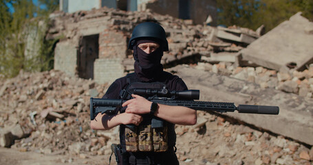 A soldier stands near a destroyed house, looks to the side and picks up his rifle