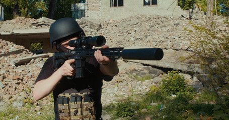 A Ukrainian soldier fires a rifle against the backdrop of a destroyed house.