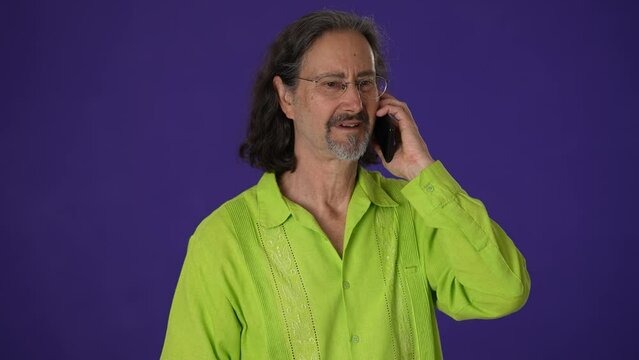 Happy excited mature man in green shirt using speaking talking on mobile cell phone just found out great big win news isolated on solid purple background studio. People lifestyle concept