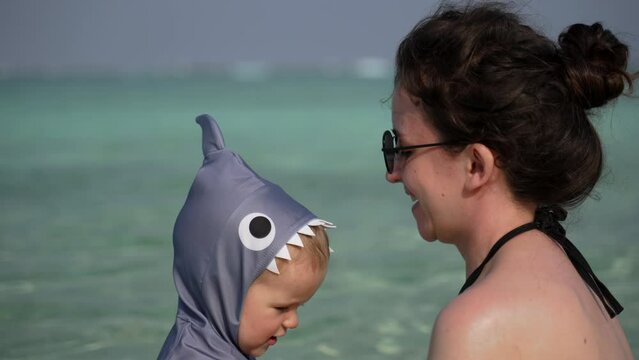 The mother raises and lowers the child in a shark suit into the ocean and back. Bathing and swimming lessons from a young age in slow motion.