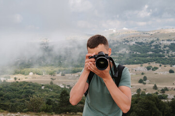 A young male tourist with a backpack climbed to the top of the mountain and takes pictures on the peak from a height. Facing the camera. The concept of active life and hiking.