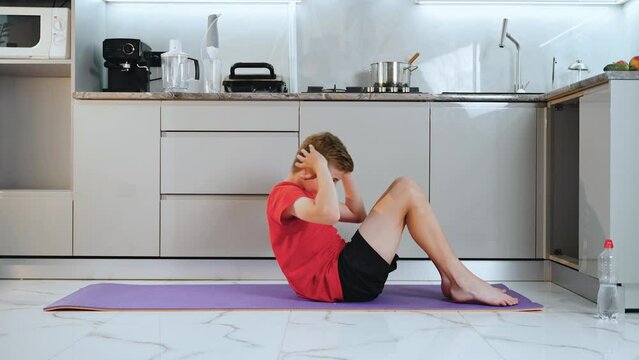 Skinny teenager quarantined in kitchen playing sports on mat. The man lies on the ground and shakes the press, tensing the abdominal muscles. The boy is training and doing exercises. Sports at home