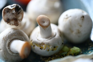 Unwashed mushrooms on a blue plate accompanied by a sliced piece of tomato with juicy seeds - close-up of the elements: stem, hat, next to the scattered lemon pepper and chopped green chives