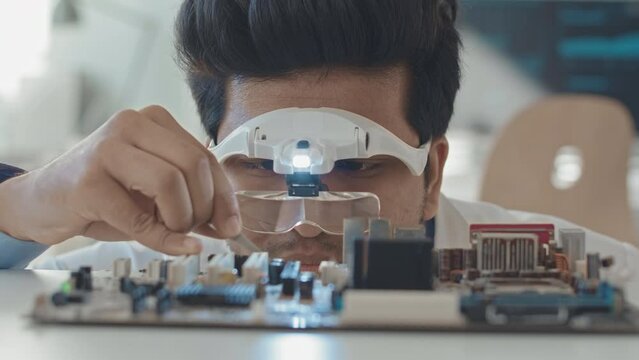 Slowmo closeup of Biracial male engineer wearing special lens loupe eyewear using tweezers while repairing motherboard in laboratory