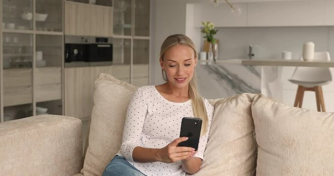 Dreamy young attractive woman editing photos in smartphone software application, communicating distantly in social network, dating online, or choosing goods in internet store, resting on sofa.