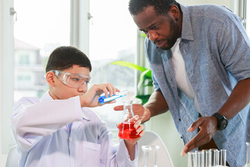A Caucasian male student raises a glass vial of various colored liquids while a teacher guides him in a science chemistry laboratory