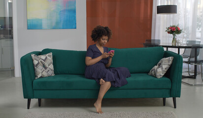 Beautiful young African-American woman in polka dot dress sits on sofa with smartphone in modern living room. Pretty female chatting with friends online at home