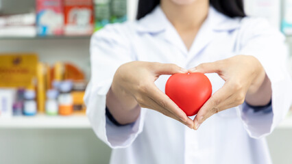 Asian female pharmacist doctor with red heart and pharmacy drug store background, medicine, pharmacy, people, health care and pharmacology.