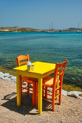 Yellow cafe restaurant table of street cafe with chairs on beach in Adamantas town on Milos island with Aegean sea with boats and yachts in background. Milos island, Greece