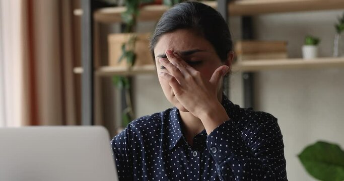 Tired young Indian ethnicity woman sit at office workplace desk in front of laptop massages nose bridge reducing headache, eye strain thinks over task feeling exhaustion after long modern tech usage