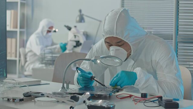 Medium slowmo of male engineer in protective coveralls, respiratory mask and gloves soldering printed circuit board in lab