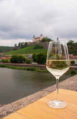 A glass of white wine standing on a wine table on an Old Bridge on Main river in front of Marienberg Fortress upon a vineyard hills in Bavarian touristic city