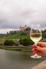 Woman hand holding a glass of white wine on an Old Bridge on Main river in front of Marienberg Fortress upon a vineyard hills in Bavarian touristic city  