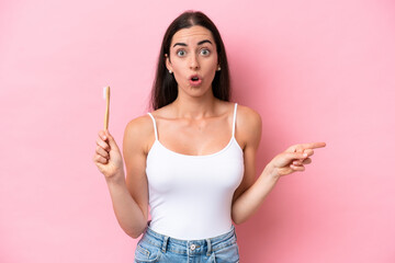 Young caucasian woman brushing teeth isolated on pink background surprised and pointing side