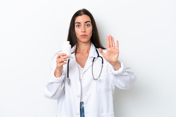 Young caucasian woman isolated on white background wearing a doctor gown and holding pills while doing stop sign