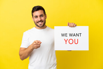 Young handsome caucasian man isolated on yellow background holding We Want You board and pointing...