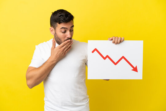 Young handsome caucasian man isolated on yellow background holding a sign with a decreasing statistics arrow symbol with surprised expression