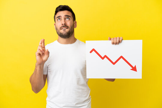 Young handsome caucasian man isolated on yellow background holding a sign with a decreasing statistics arrow symbol with fingers crossing