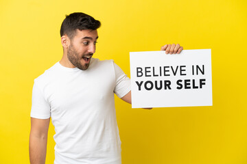 Young handsome caucasian man isolated on yellow background holding a placard with text Believe In Your Self