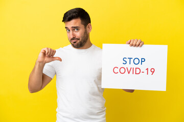 Young handsome caucasian man isolated on yellow background holding a placard with text Stop Covid 19 with proud gesture