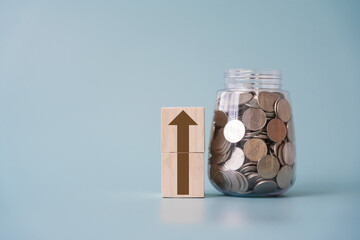 Brown arrow going up on wooden blocks beside Thai currency coins jar and light blue background. Concept of growing or improving in positive direction.