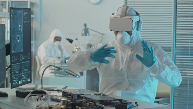 Medium slowmo of unrecognizable electronics engineer in protective coveralls and respiratory mask using VR headset in lab while working with computer components