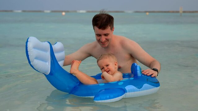Father and son wave at the camera smiling and playing in the water with a rubber toy. Toddler rejoices when Dad spins around his axis rubber toy