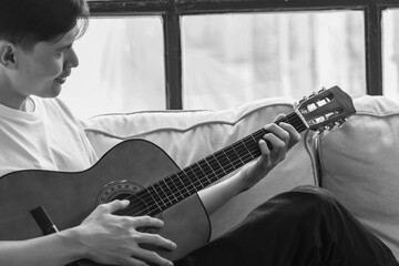 Young asian man playing guitar while sitting on sofa in living room Black and white He love playing...