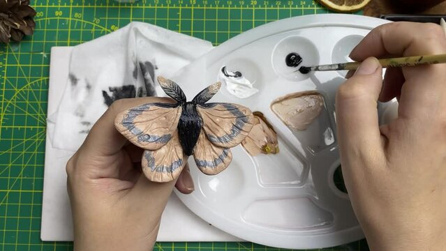 Female's hands paint acrylic brooch in form of butterfly. Design of jewelry in form of moth made of clay. Palette, brushes, napkins on wooden table. Handmade and craft on quarantine.