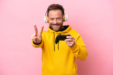 Middle age man playing with a video game controller isolated on pink background smiling and showing...