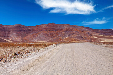 Fototapeta na wymiar Dirt road in arid barren dry landscape, red mountains, clear blue sky - Road trip to Punta de Jandia, Fuerteventura