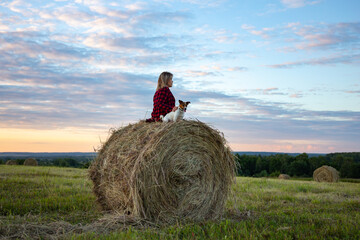 Naklejka na ściany i meble Young woman with her little dog jack russell terrier sitting on a haystack in a field on a summer evening at sunset