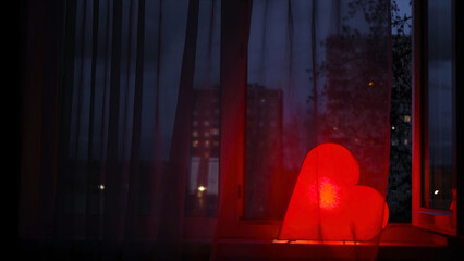 Bright red heart shines at window at night. Concept. Night light in shape of heart stands on windowsill and shines brightly for lovers. Love and Valentine's day