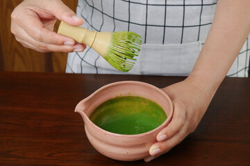 Hand hold whisk preparing and mixing traditional organic Japanese matcha green tea powder with milk in pink bowl on woo table