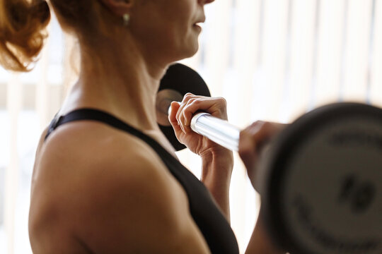 Side View Of Fit Adult Slim Woman Lifting Barbell In Fitness Box. Close-up On The Arm With Muscles In Front Of The Window. Workout At Home Or Outdoors