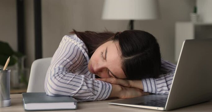 Overworked female office employee sleeps put head on arms lying at workplace desk near laptop, close up. Bored student take break fall asleep instead of studying. Fatigue, unmotivated worker concept