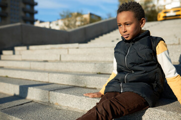Adorable little boy sitting on stairs in city, wearing velvet jeans and black vest, carrying...