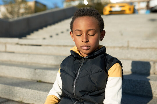 Thoughtful African schoolboy sitting on concrete stairs outdoors, dressed in urban style, carrying bag on shoulders, looking down, isolated against cityscape background with parked cars - Powered by Adobe
