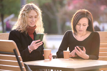 Two distracted female friends browsing their mobile phones while sitting together at city street cafe. Girlfriends ignoring each other while staring in gadgets. Addiction from social media concept