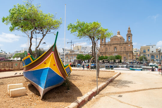 A traditional Maltese fishing boat called a Luzzu on land near the marina. In the background is the Parish church of Saint Joseph.