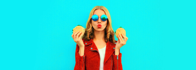 Portrait of stylish young woman with tasty big burger fast food blowing her lips on blue...