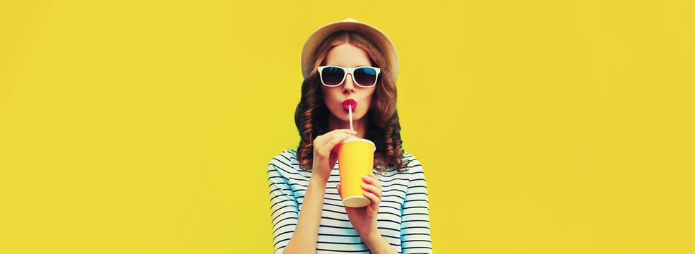 Portrait of stylish young woman drinking fresh juice wearing summer straw hat, sunglasses and striped t-shirt on yellow background