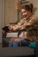 A gamer girl in jeans and a plaid shirt emotionally plays a video game on a portable game console at home, in the living room. Online games with friends, youth culture.
