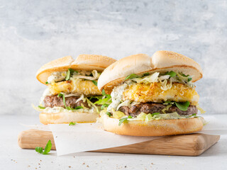 Burger with mozarella fried in batter, beef cutlet, rocket salad, lettuce and creamy sauce. Burgers on wooden cutting board. Close up view. Homemade tasty snack, fast food or lunch. Grey background.