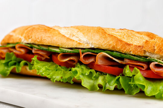 Close up baguette with ham and vegetables. Sandwich with ham, tomato, lettuce salad, cucumber on marble board.
